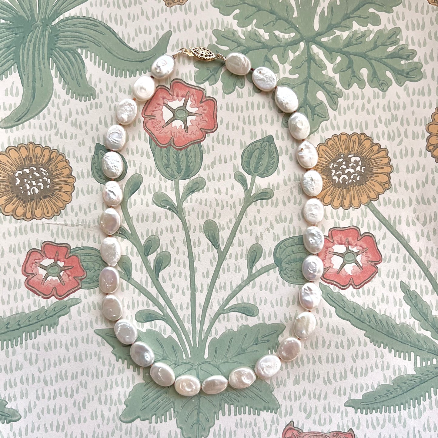 A knotted silk oval pearl necklace with a gold-fill fish hook clasp, displayed against a floral patterned background.