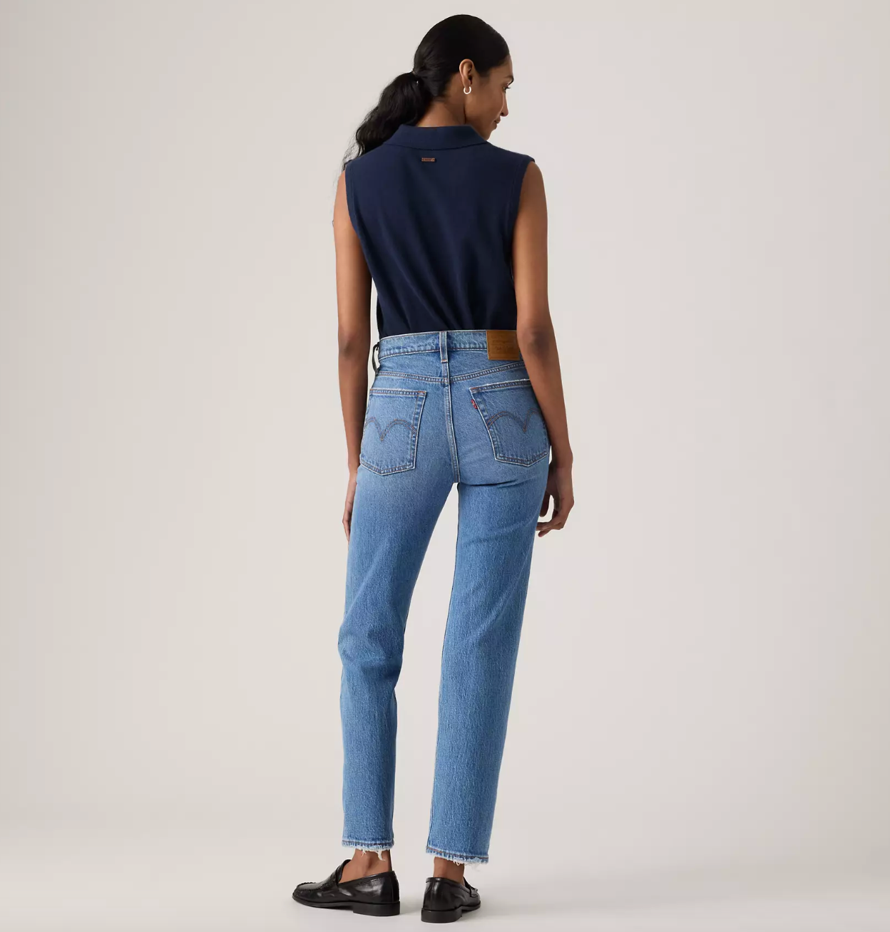 A model is shown from the back wearing a navy sleeveless collared top, light blue Levi's jeans with the brand's signature heart patch on the back pockets, and black leather loafers, against a plain light gray studio background.