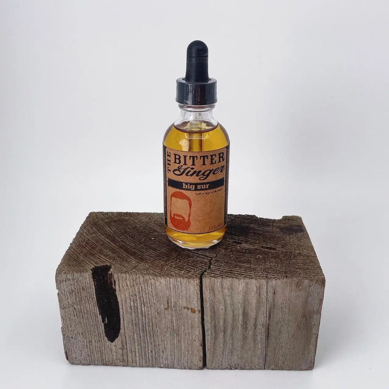 The Bitter Ginger Big Sur Bitters 2 oz glass dropper bottle with kraft paper label, displayed upright on a rustic weathered wood block against a white background