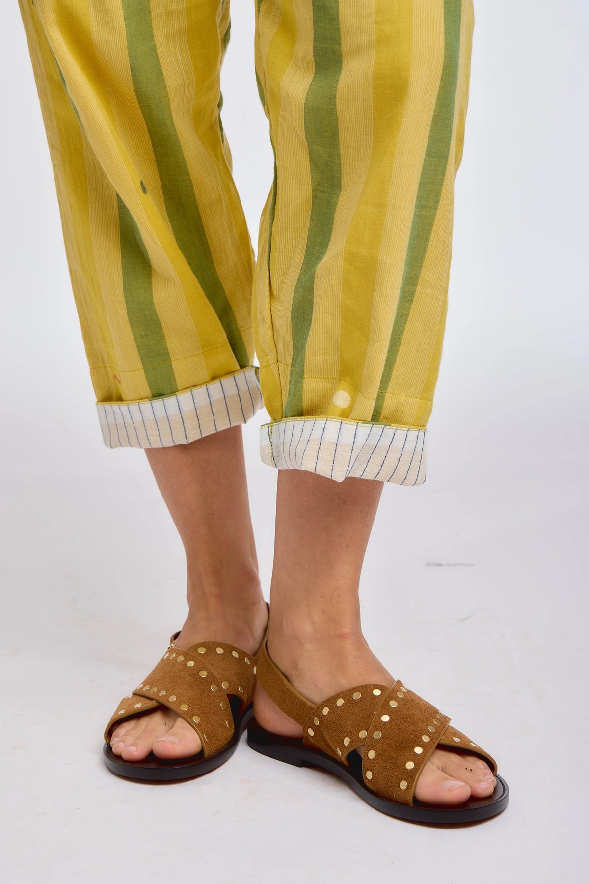 Close-up of DVAA Celeste Pant in Goldenrod hem and footwear detail against white studio backdrop showing goldenrod yellow and sage green wide vertical stripe handwoven cotton, cropped tapered leg with contrast white pinstripe cuff turn-up, styled with tan leather gold-studded crossband slide sandals