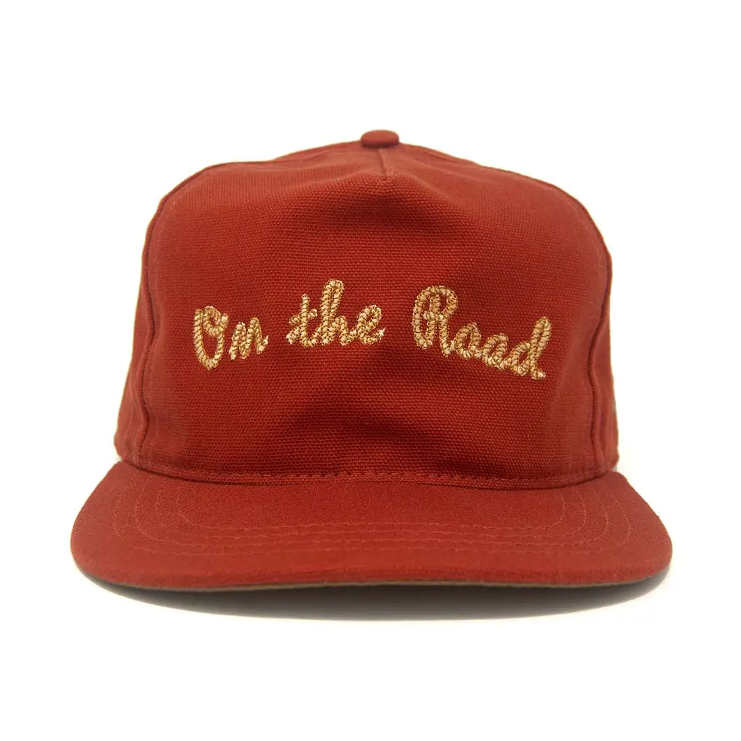 A rust red snapback cap with 'On the Road' embroidered in gold cursive on the front, set against a plain white background.