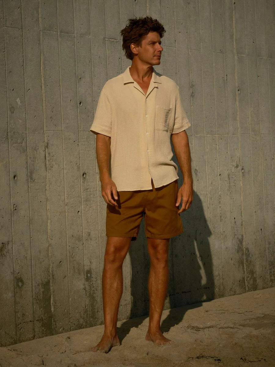 Man wearing a beige shirt and brown shorts standing against a concrete wall.