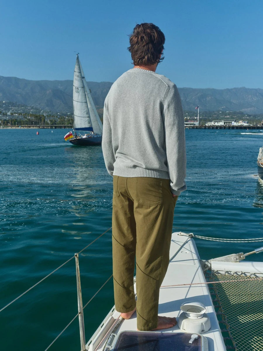 Back view of a man standing on the bow of a sailboat wearing the Mollusk Hemp Cambridge Sweater in Gray Flannel with olive trousers, overlooking open water with a sailboat and coastal mountains in the distance