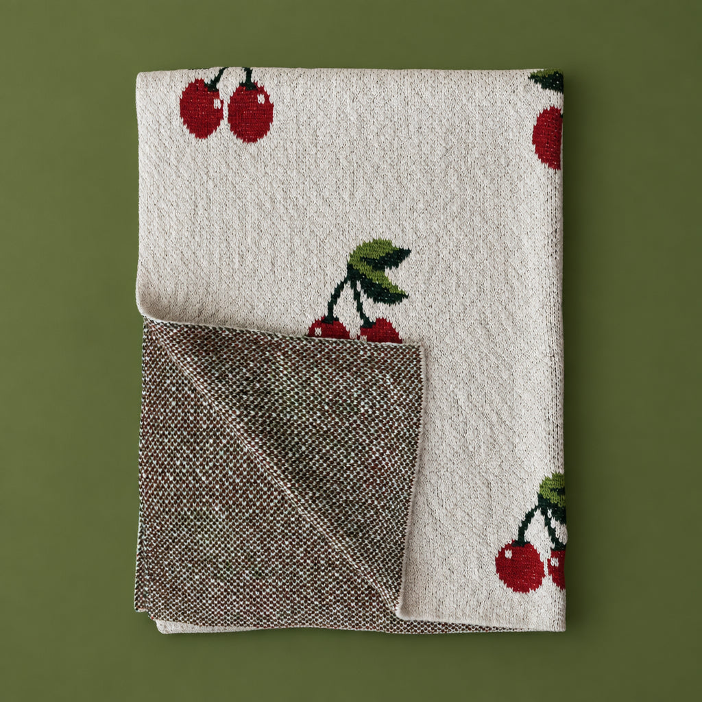 Folded view of the Dotter Cotton Knit Baby Blanket with Cherries, showing the cream cherry-print knit face and the textured olive and brown reverse side, on a sage green background