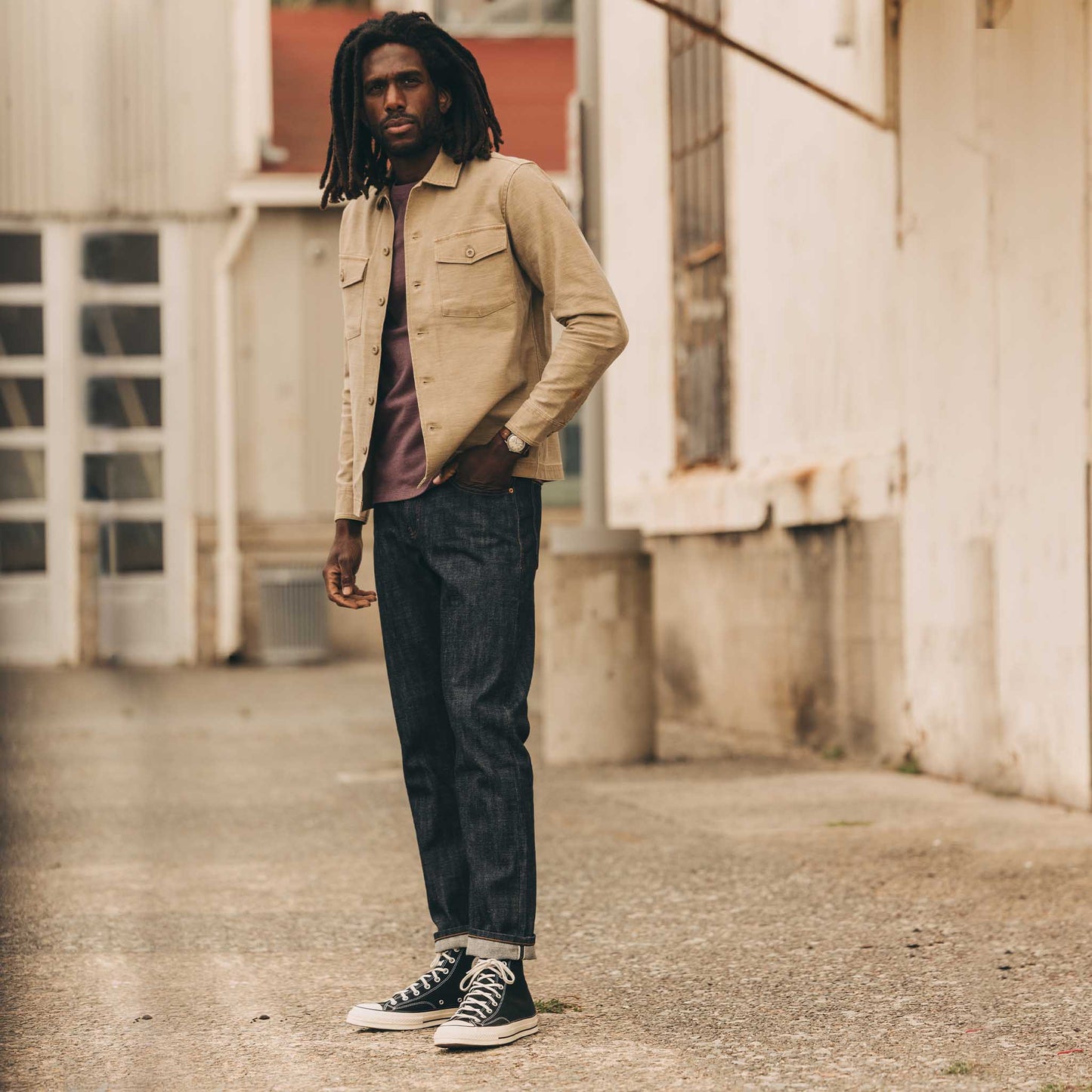 Full-length lifestyle shot of a man wearing The Democratic Jean in Rigid Organic Selvedge by Taylor Stitch with a tan overshirt and black Converse, standing in an urban alleyway