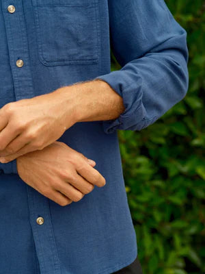 Close-up of Mollusk Surf Shop One Pocket Shirt in Indigo Flannel showing tonal corozo-style button-front placket, single left-chest patch pocket, rolled long sleeve with button cuff, and soft indigo blue 80% cotton 20% linen flannel fabric texture against lush green backdrop