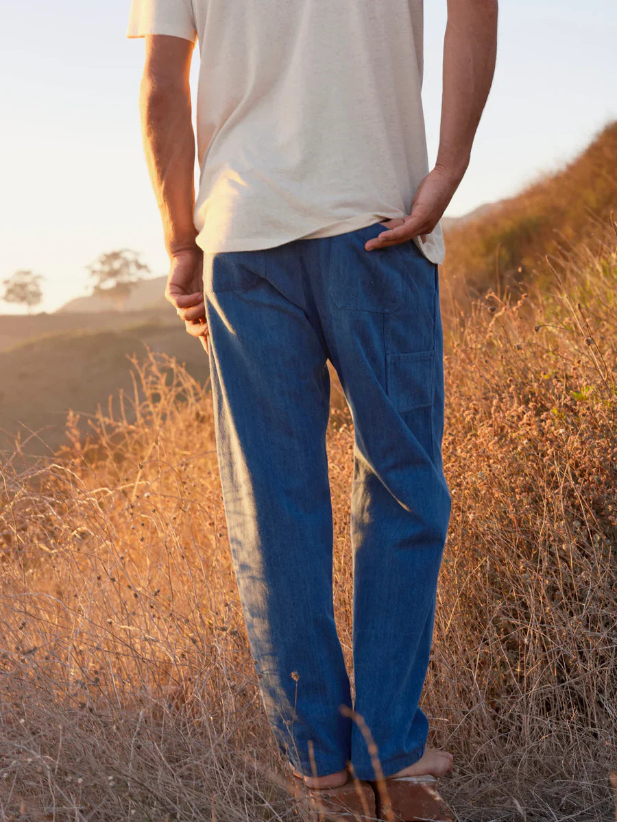 Back view of male model wearing medium indigo denim relaxed straight-leg pants, showing the elastic waistband, clean back panel, and slight leg taper, styled with a white t-shirt and sandals in a golden hillside meadow at dusk.