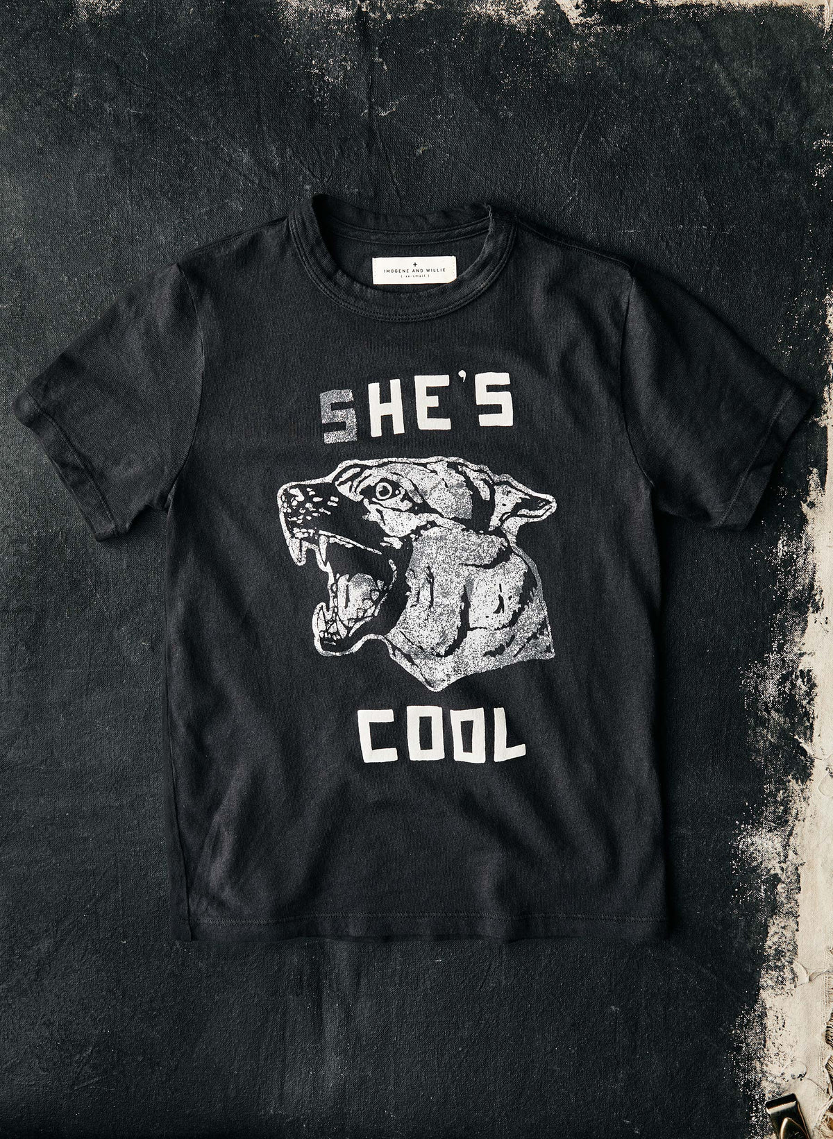 The She's Cool Tee by Imogene + Willie laid flat on a dark textured surface, showing the full front with a vintage-style screen-printed graphic of a snarling panther head and the text 'SHE'S COOL' in white distressed lettering on a black cotton tee