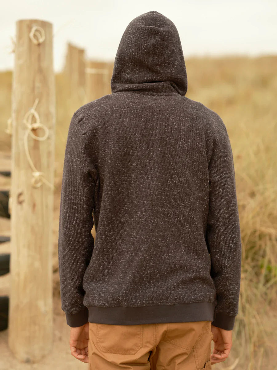 Back view of male model wearing a faded navy garment-dyed zip-up hoodie with hood up, showing the textured terry knit fabric, relaxed fit, and ribbed hem and cuffs, styled with tan pants against a coastal dune background.