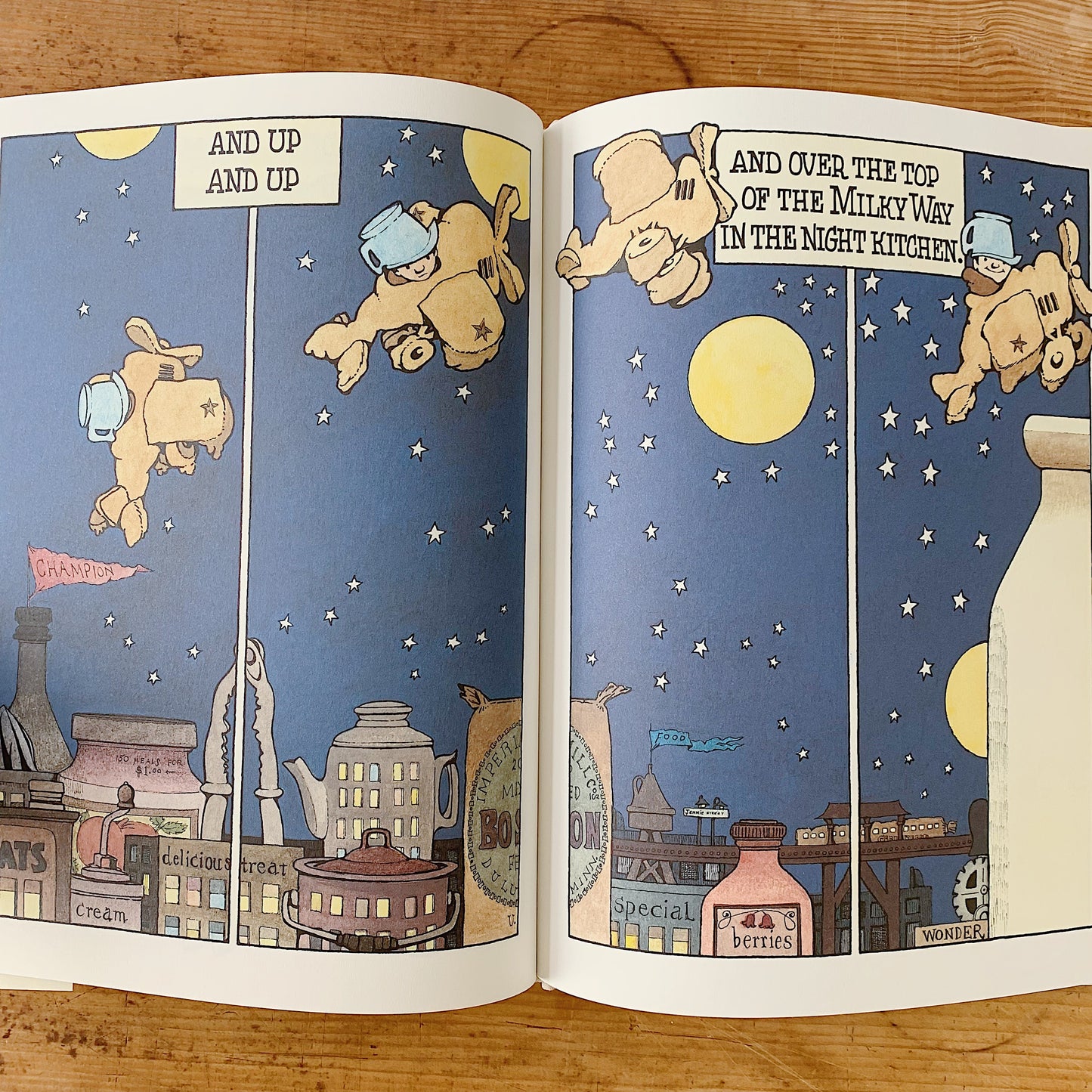 Open interior spread of In the Night Kitchen showing a four-panel comic-strip sequence across both pages: Mickey — wearing a blue chef's hat and molded into a bread-dough airplane shape — flies upward through a deep blue star-studded night sky past a full yellow moon, with caption banners reading "AND UP AND UP" on the left and "AND OVER THE TOP OF THE MILKY WAY IN THE NIGHT KITCHEN!" on the right. Below, a whimsical cityscape of oversized food containers serves as buildings, including jars labeled Cream, O