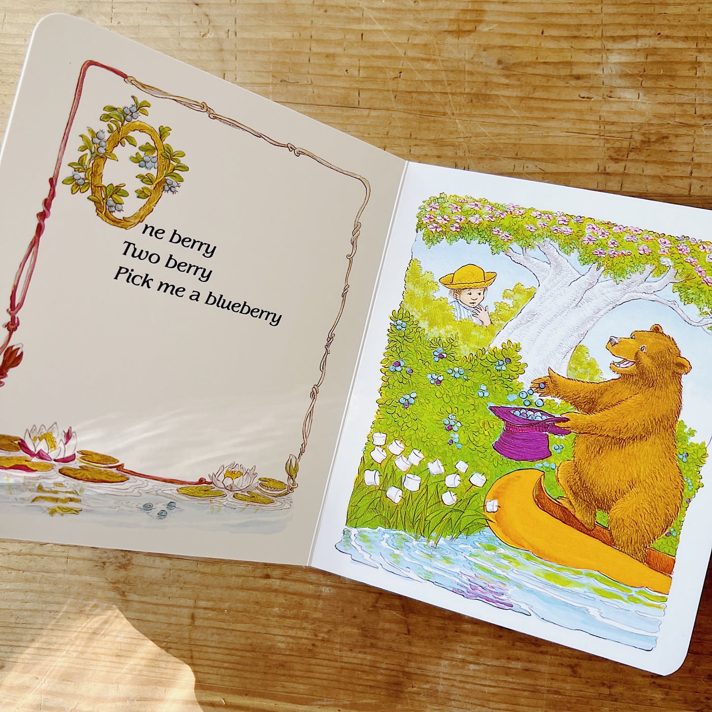 Open interior spread of Jamberry board book: on the left, a cream page with a decorative twig-and-berry border and an ornate illuminated capital O entwined with blueberry branches, with the verse "One berry / Two berry / Pick me a blueberry" in black serif type, and a lily-pad pond scene along the bottom edge; on the right, a full-color illustration of a large brown bear sitting at the edge of a stream, filling a purple top hat with blueberries, while a small boy in a yellow hat peeks down from a flowering