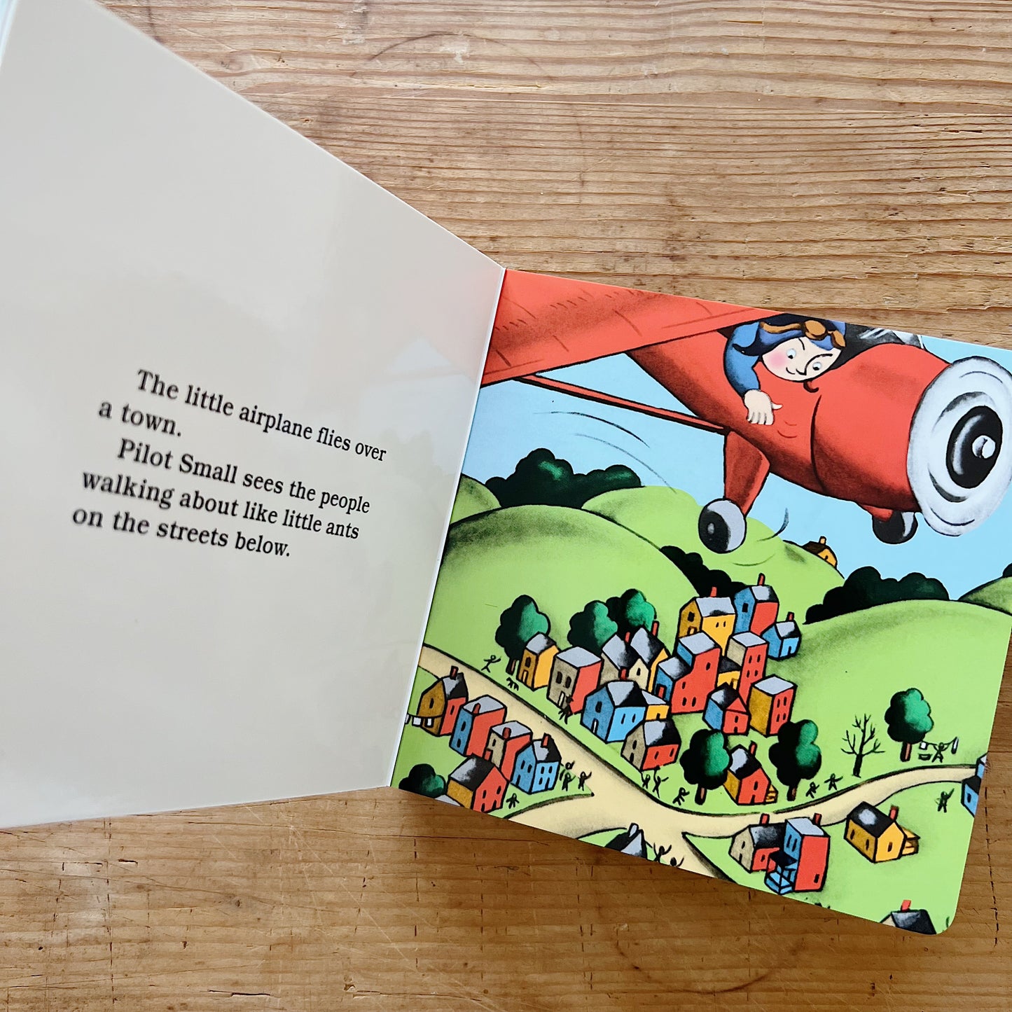 Interior spread of The Little Airplane showing Pilot Small leaning out of his red airplane as it flies over a colorful town below, beside text reading "The little airplane flies over a town. Pilot Small sees the people walking about like little ants on the streets below"