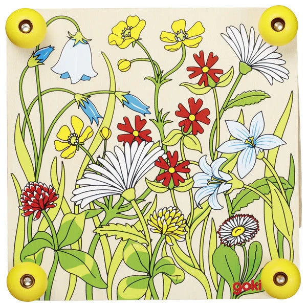 Top view of the Goki Flower Press wooden plate showing a colorful illustrated wildflower meadow with daisies, bluebells, buttercups, clover, and other flowers, with four yellow screw knobs at the corners and the Goki logo in the bottom right