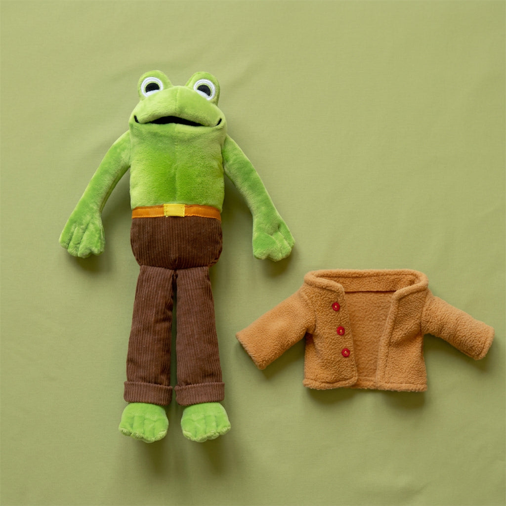 Green Frog plush toy in brown corduroy pants, lying beside its removed camel-colored coat with red buttons.