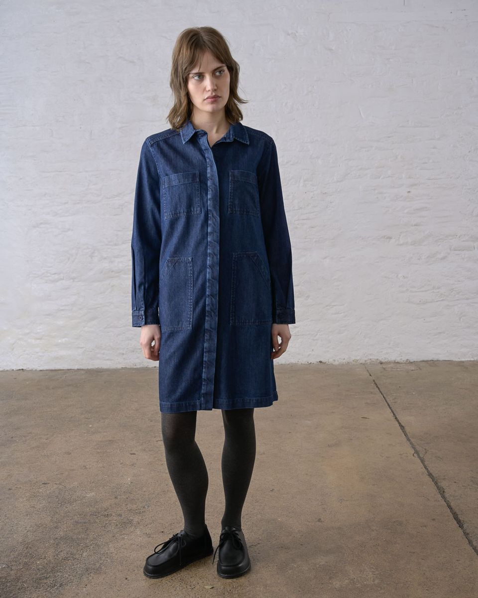 Full-length front view of a woman wearing the BIBICO Ocean Shirt Dress in Denim buttoned closed, showing the pointed collar, concealed button placket, two chest patch pockets, two large hip patch pockets, buttoned cuffs, and above-the-knee length in deep indigo 9oz cotton denim, styled with dark tights and black lace-up shoes against a whitewashed brick wall
