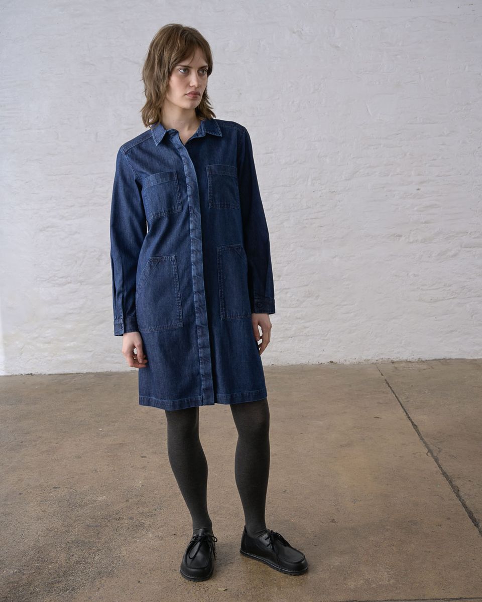 Three-quarter front view of a woman wearing the BIBICO Ocean Shirt Dress in Denim, showing the pointed collar, concealed button front, chest and hip patch pockets, relaxed boxy silhouette, and above-the-knee hem in deep indigo 9oz cotton denim, styled with dark tights and black lace-up shoes against a whitewashed brick wall
