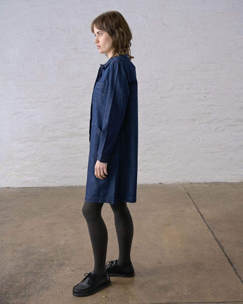Pure side profile of a woman wearing the BIBICO Ocean Shirt Dress in Denim, showing the relaxed boxy silhouette, full-length sleeve with buttoned cuff, pointed collar, and above-the-knee hem in deep indigo 9oz cotton denim, styled with dark tights and black lace-up shoes against a whitewashed brick wall
