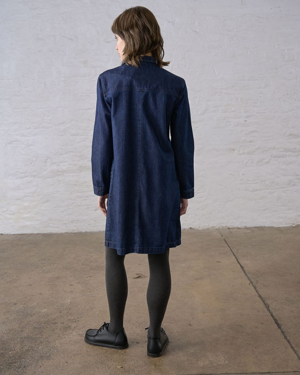 Back view of a woman wearing the BIBICO Ocean Shirt Dress in Denim, showing the back yoke, center back seam, relaxed boxy silhouette, full-length sleeves with buttoned cuffs, and above-the-knee hem in deep indigo 9oz cotton denim, styled with dark tights and black lace-up shoes against a whitewashed brick wall

