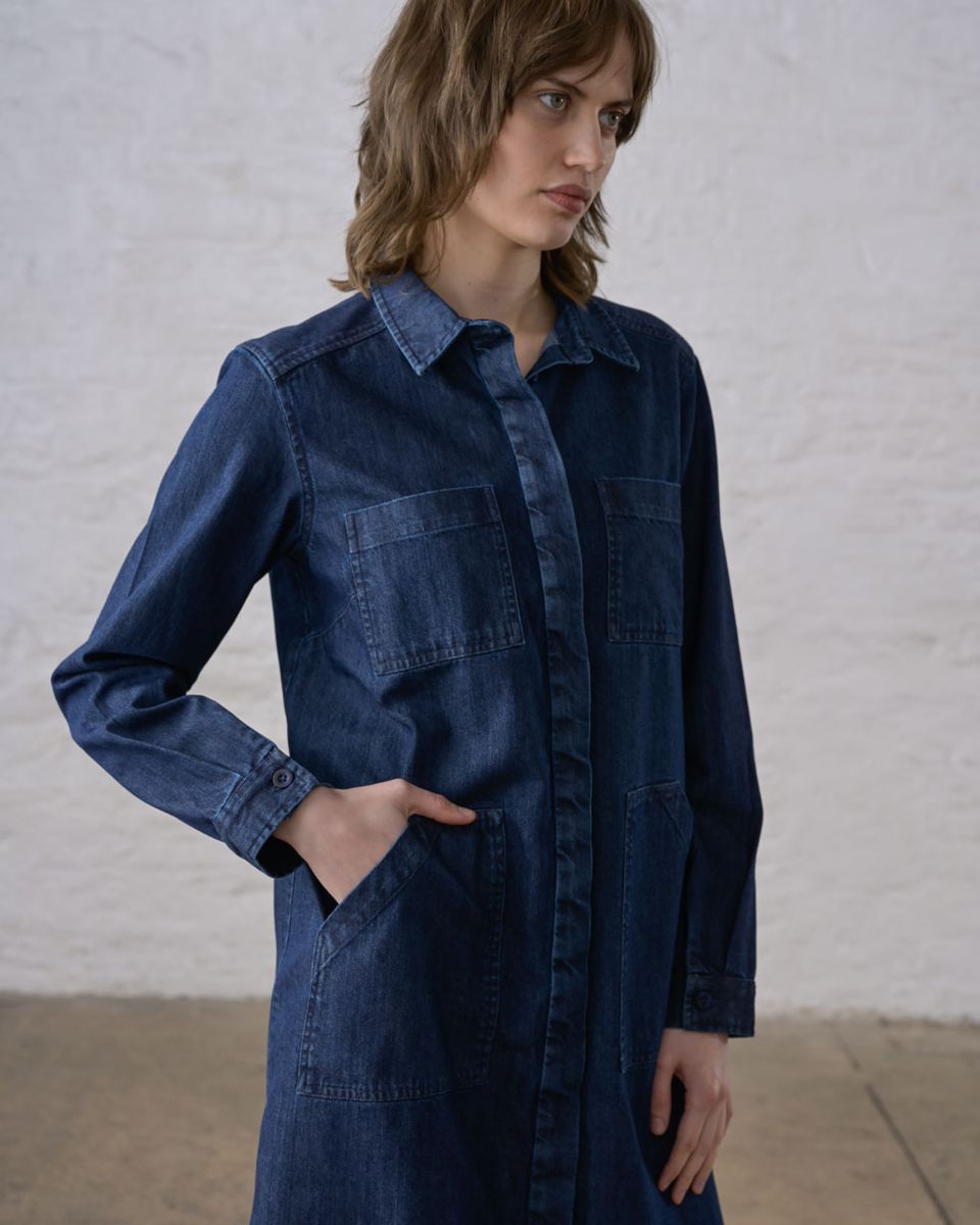 Close three-quarter front view of a woman wearing the BIBICO Ocean Shirt Dress in Denim with hand in the hip pocket, showing the pointed collar, two chest patch pockets, deep carpenter-style hip pocket, concealed button placket, and buttoned cuff detail in deep indigo 9oz cotton denim, against a whitewashed brick wall

