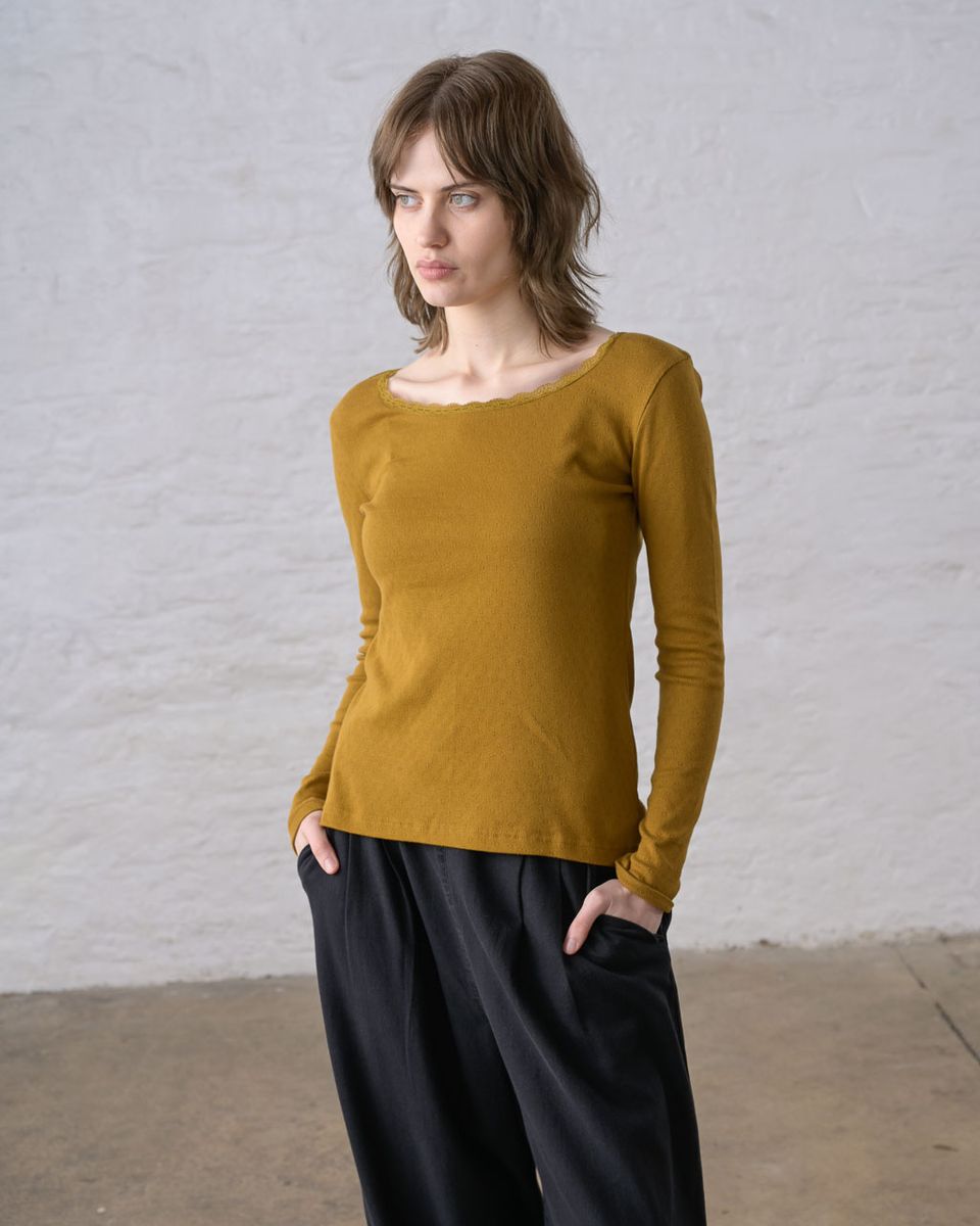 Three-quarter front view of a woman wearing the BIBICO Cali Pointelle Tee in Bronze, showing the scooped neckline with scalloped lace trim, fitted silhouette, long sleeves, and pointelle-textured organic cotton in rich bronze-mustard gold, styled with black wide-leg pants against a whitewashed brick wall
