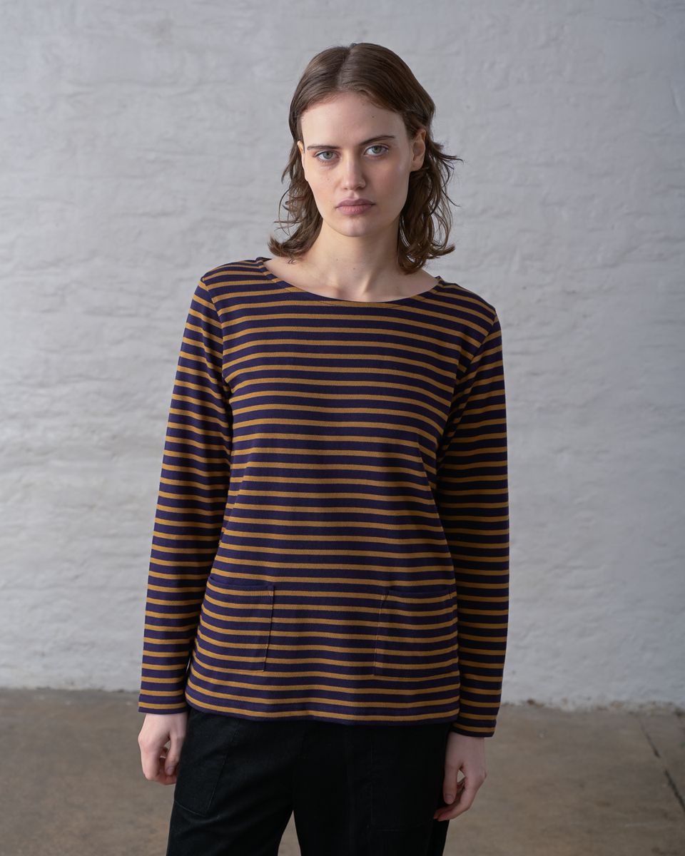 Front portrait of a woman wearing the BIBICO Long Sleeve Sailor Tee in Bronze and Navy Stripe, showing the wide boat neckline, relaxed fit, and custom-dyed horizontal stripe in mid-weight organic cotton jersey, styled with black wide-leg pants against a whitewashed brick wall
