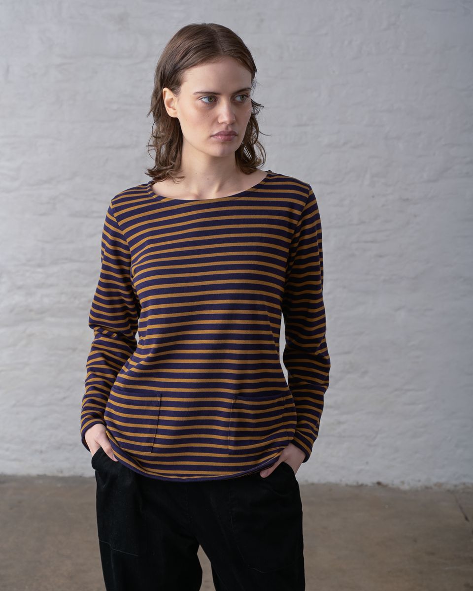 Three-quarter view of a woman looking to the side wearing the BIBICO Long Sleeve Sailor Tee in Bronze and Navy Stripe, showing the relaxed fit, wrist-length sleeves, curved hem, and custom-dyed horizontal stripe in organic cotton jersey, styled with black wide-leg pants against a whitewashed brick wall
