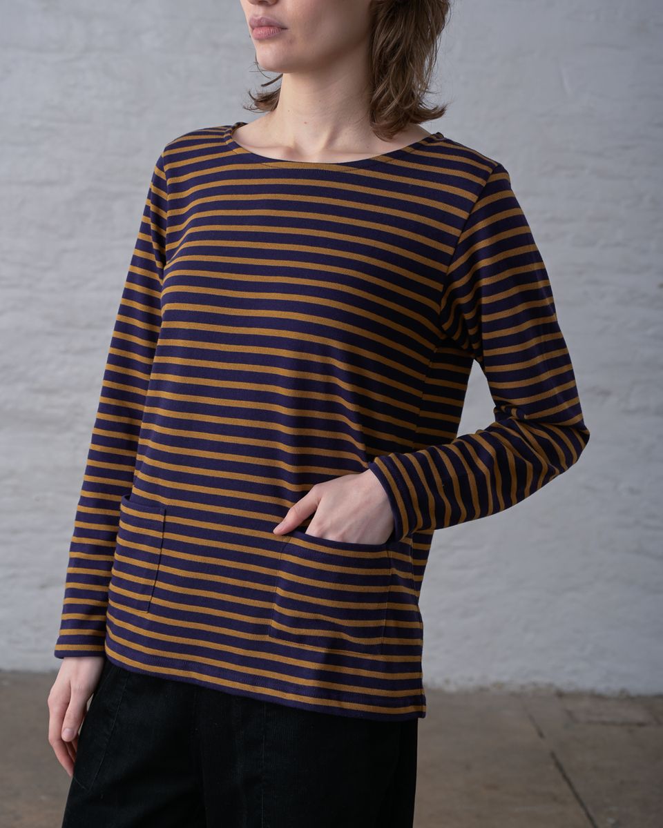 Three-quarter front view of a woman wearing the BIBICO Long Sleeve Sailor Tee in Bronze and Navy Stripe with hand in front patch pocket, showing the wide boat neckline, relaxed silhouette, two front patch pockets, and custom-dyed horizontal stripe in organic cotton jersey, styled with black wide-leg pants against a whitewashed brick wall
