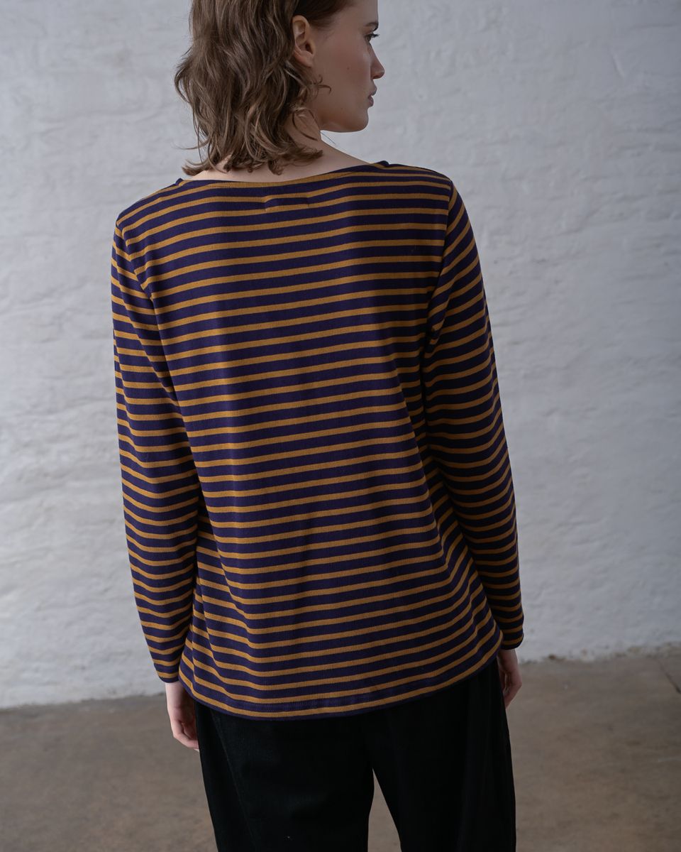 Back view of a woman wearing the BIBICO Long Sleeve Sailor Tee in Bronze and Navy Stripe, showing the wide boat back neckline, relaxed silhouette, wrist-length sleeves, and custom-dyed bronze and navy horizontal stripe in organic cotton jersey, styled with black wide-leg pants against a whitewashed brick wall
