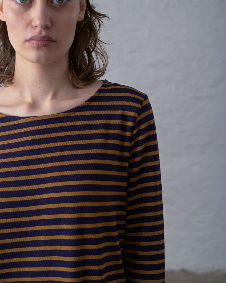 Close cropped front detail of a woman wearing the BIBICO Long Sleeve Sailor Tee in Bronze and Navy Stripe, showing the wide boat neckline, shoulder seam, and the custom-dyed bronze and navy horizontal stripe texture of the mid-weight organic cotton jersey, against a whitewashed brick wall
