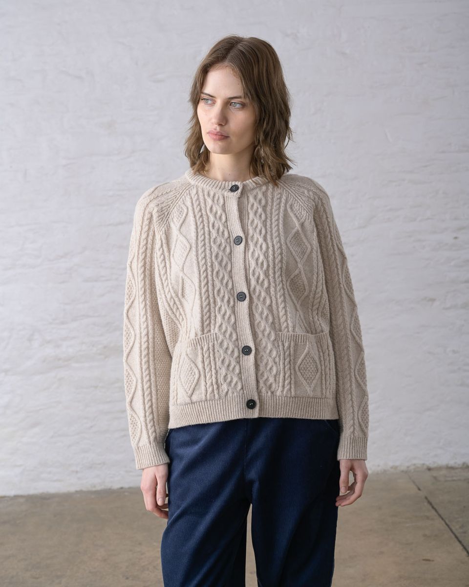 Front view of a woman wearing the BIBICO Derby Aran Cardigan in Oat, showing the crew neck with ribbed trim, imitation corozo button front, all-over Aran cable knit in diamond and rope panels, two front patch pockets, and raglan sleeves in warm oat lambswool, styled with navy wide-leg pants against a whitewashed brick wall

