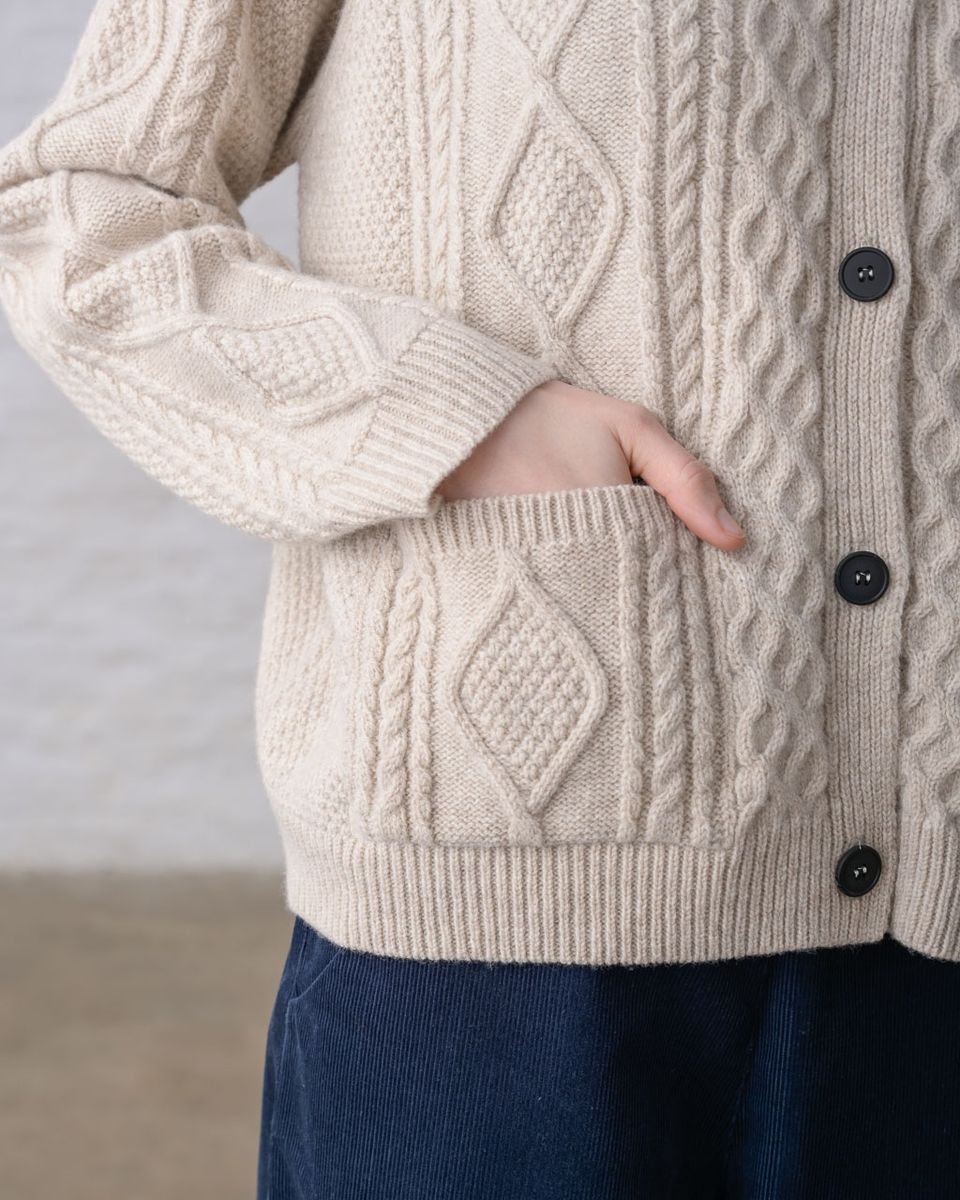 Close detail of the hip and front pocket of the BIBICO Derby Aran Cardigan in Oat, showing a hand in the cable-knit patch pocket with ribbed trim, button front with imitation corozo buttons, ribbed hem, and the Aran cable knit texture in oat lambswool, styled over navy corduroy pants
