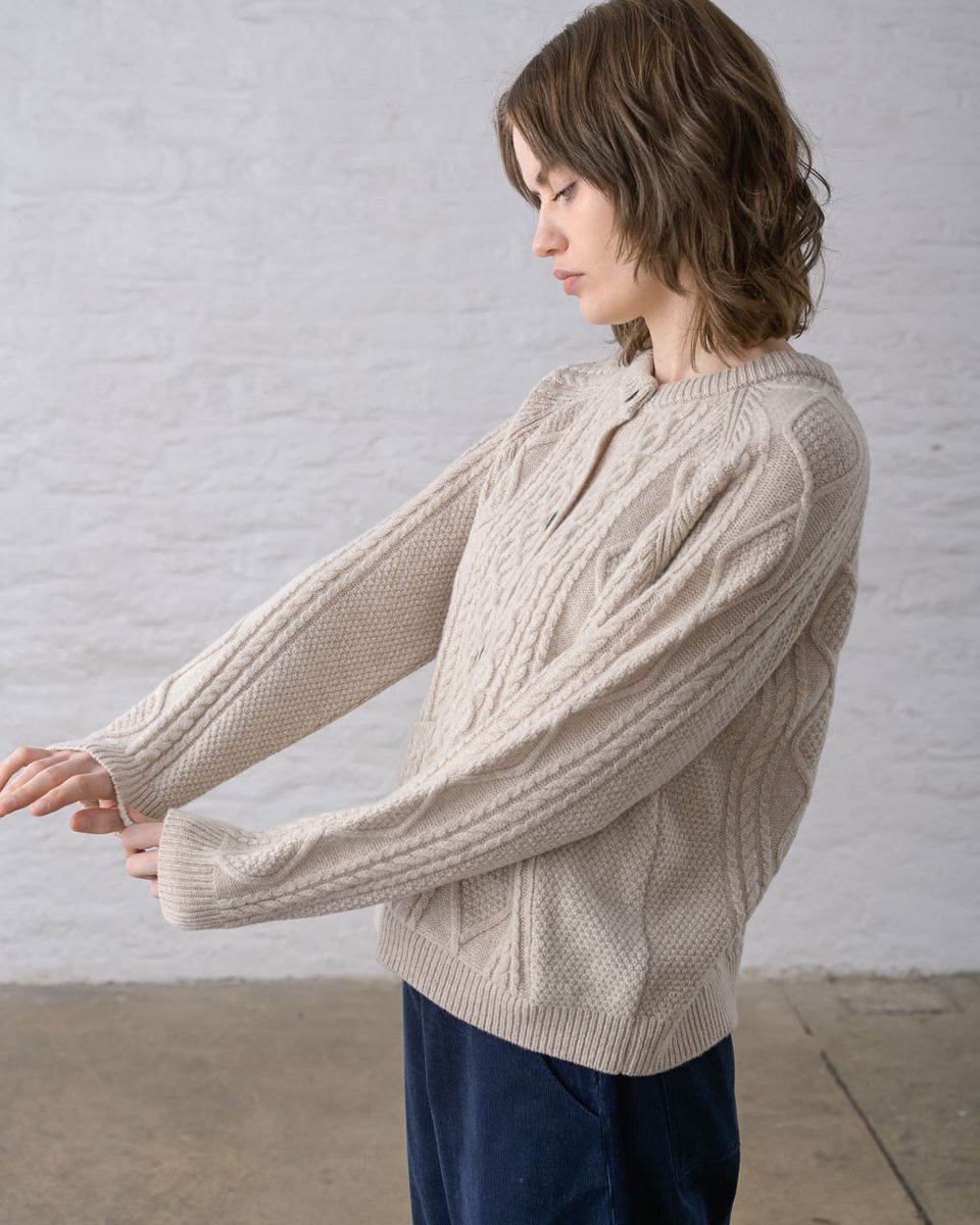 Pure side profile of a woman holding the cuff of the BIBICO Derby Aran Cardigan in Oat, showing the raglan sleeve, ribbed cuff, all-over Aran cable knit texture, and relaxed silhouette in warm oat lambswool, styled with navy wide-leg pants against a whitewashed brick wall
