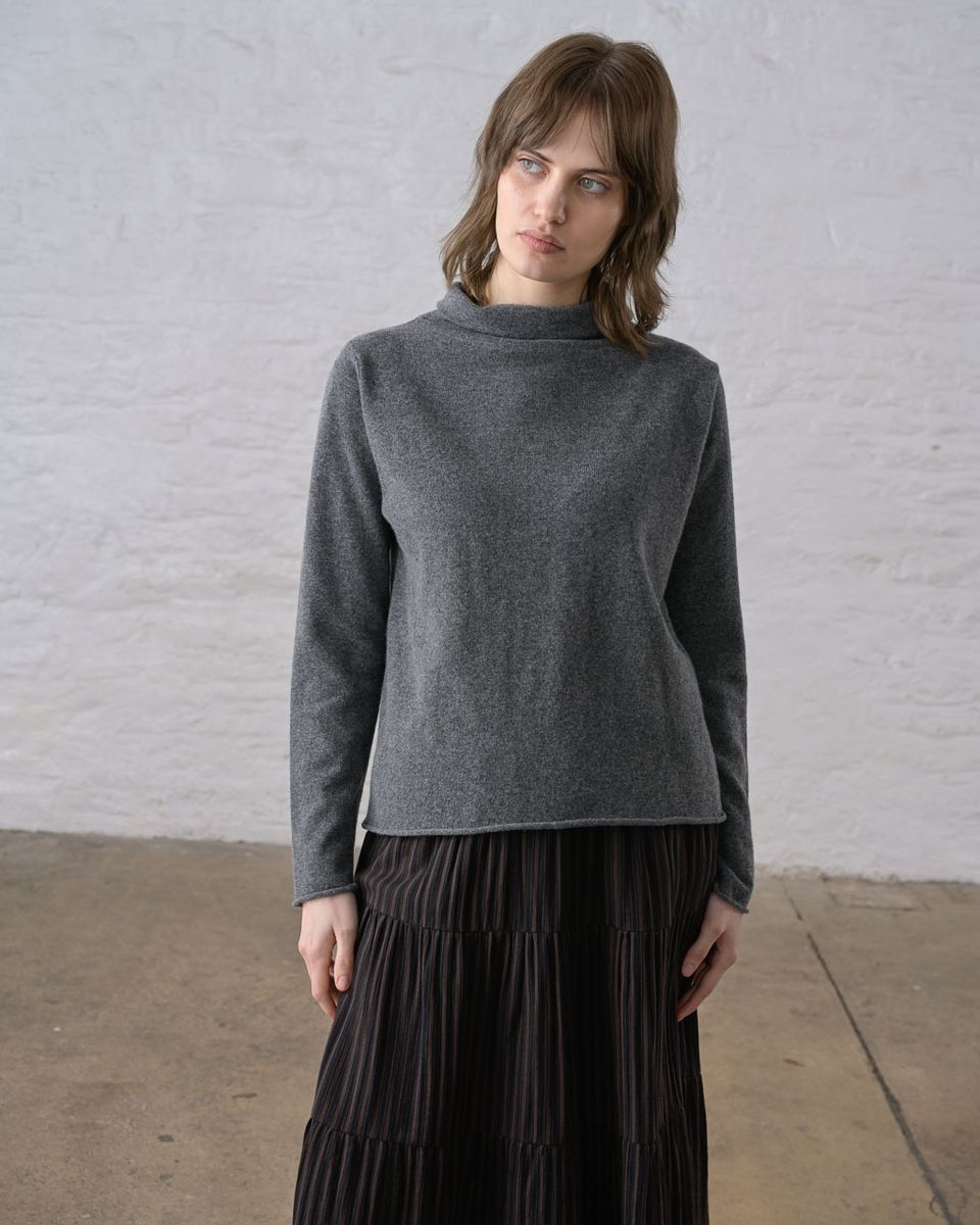 Front view of a woman wearing the BIBICO Aria Jumper in Grey Marle, showing the short roll-up funnel neck, subtle A-line silhouette, long sleeves, rolled hem, and fine pointelle-textured knit in lightweight grey marle Geelong Merino wool, styled with a dark pleated midi skirt against a whitewashed brick wall
