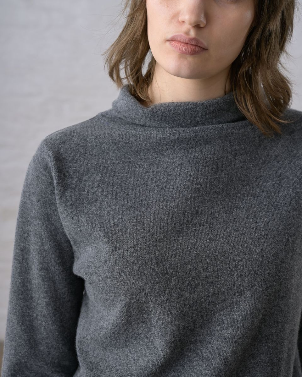 Close upper-body detail of a woman wearing the BIBICO Aria Jumper in Grey Marle, showing the short roll-up funnel neck, fine pointelle-textured knit, and the heathered grey marle tone of the lightweight Geelong Merino wool against a whitewashed brick wall
