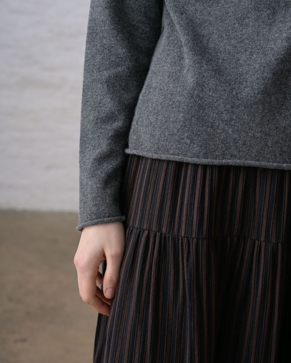 Close lower-body detail of a woman wearing the BIBICO Aria Jumper in Grey Marle, showing the rolled hem, long sleeve with rolled cuff, and the fine knit texture in lightweight grey marle Geelong Merino wool, styled over a dark pleated tiered midi skirt
