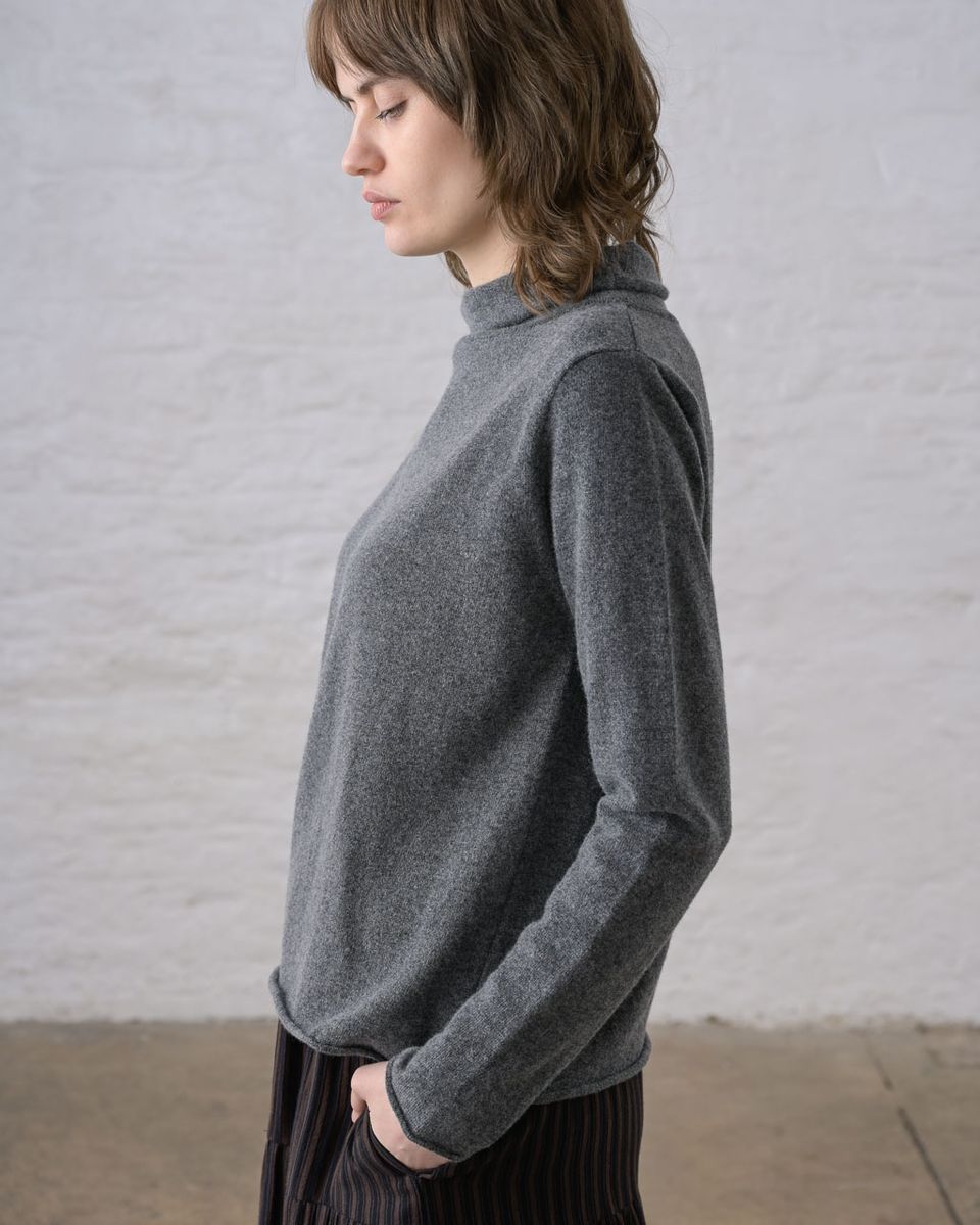 Side profile of a woman wearing the BIBICO Aria Jumper in Grey Marle with eyes downcast, showing the short funnel neck, A-line silhouette, long sleeve with rolled cuff, and fine knit texture in lightweight grey marle Geelong Merino wool, styled with a dark pleated midi skirt against a whitewashed brick wall
