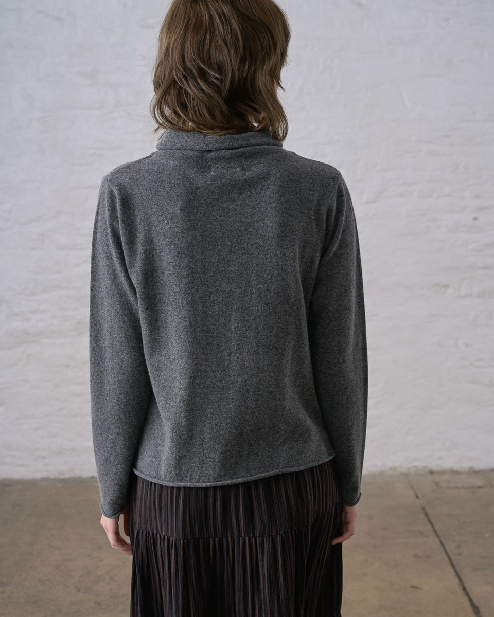 Back view of a woman wearing the BIBICO Aria Jumper in Grey Marle, showing the short funnel neck, A-line shape, long sleeves, rolled hem, and fine pointelle-textured knit in lightweight grey marle Geelong Merino wool, styled with a dark pleated midi skirt against a whitewashed brick wall
