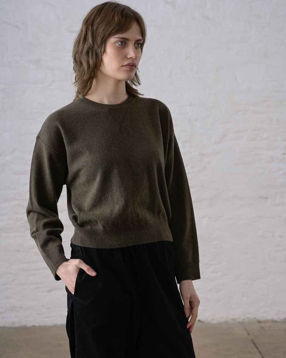 Three-quarter front view of a woman wearing the BIBICO Ona Jumper in Olive, showing the ribbed crew neckline, dropped shoulders, wide long sleeves, wide ribbed hem band, and cropped silhouette in lightweight olive Geelong Merino wool, styled with black wide-leg trousers against a whitewashed brick wall
