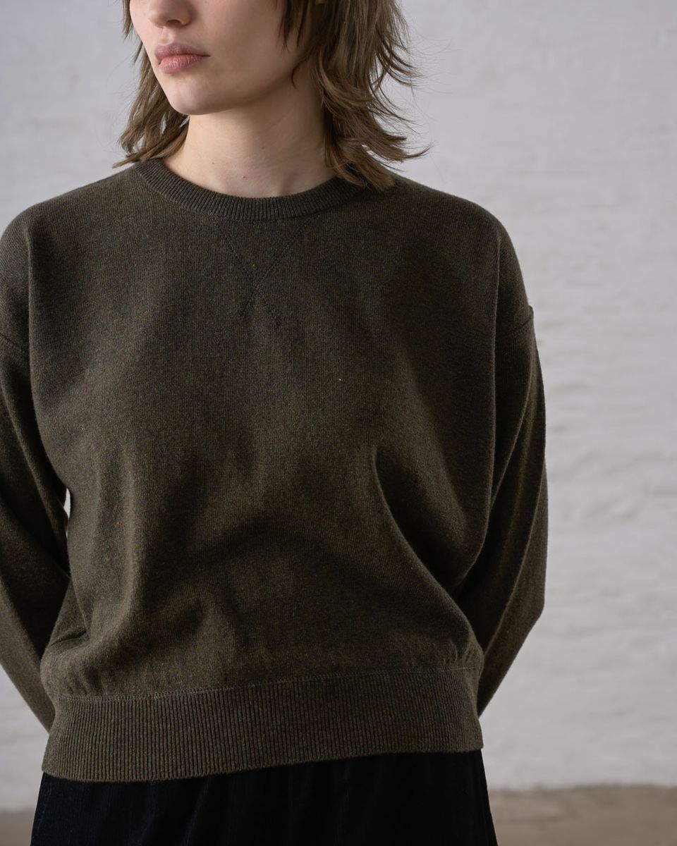 Close front detail of a woman wearing the BIBICO Ona Jumper in Olive, showing the ribbed crew neckline, dropped shoulder seam, long sleeves with ribbed cuffs, wide ribbed hem band, and the fine smooth knit texture in lightweight olive Geelong Merino wool, against a whitewashed brick wall
