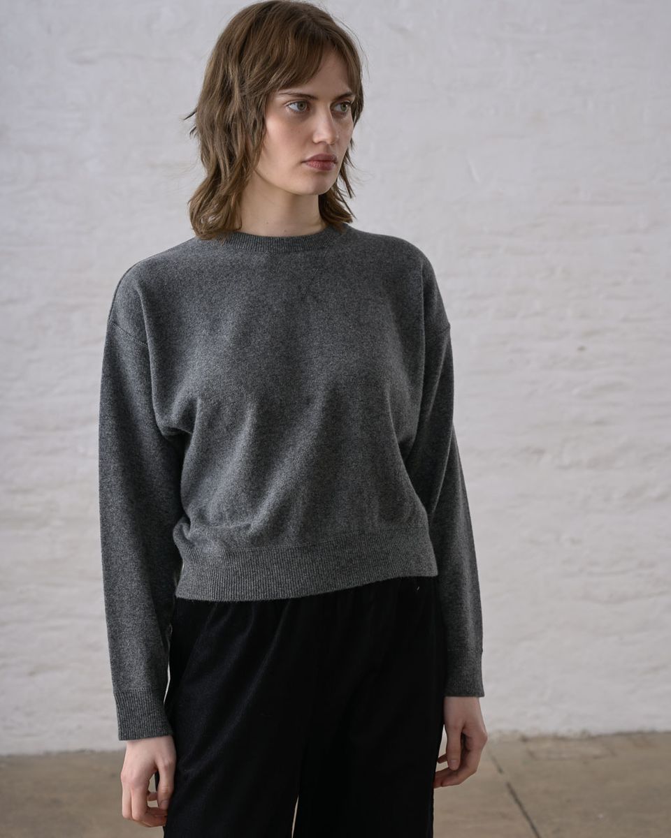 Three-quarter front view of a woman wearing the BIBICO Ona Jumper in Grey Marle, showing the ribbed crew neckline, dropped shoulders, wide long sleeves, wide ribbed hem band, and cropped silhouette in lightweight heathered grey marle Geelong Merino wool, styled with black wide-leg trousers against a whitewashed brick wall
