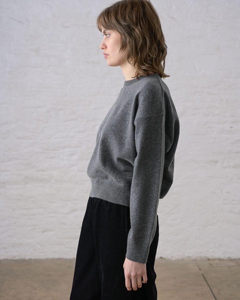 Pure side profile of a woman wearing the BIBICO Ona Jumper in Grey Marle, showing the dropped shoulder, wide long sleeve with ribbed cuff, wide ribbed hem band, and the cropped silhouette in lightweight heathered grey marle Geelong Merino wool, styled with black wide-leg trousers against a whitewashed brick wall
