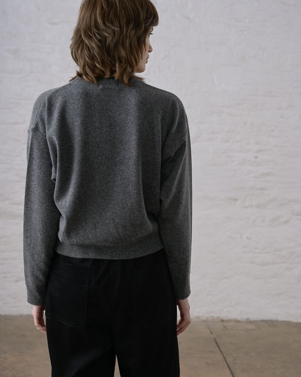 Back view of a woman wearing the BIBICO Ona Jumper in Grey Marle, showing the dropped shoulders, wide long sleeves with ribbed cuffs, wide ribbed hem band, and cropped silhouette in lightweight heathered grey marle Geelong Merino wool, styled with black wide-leg trousers against a whitewashed brick wall
