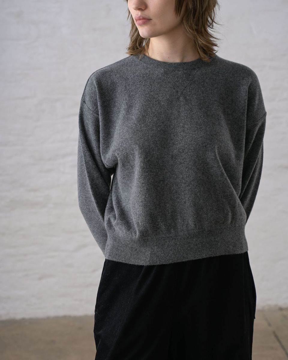 Close front detail of a woman wearing the BIBICO Ona Jumper in Grey Marle, showing the ribbed crew neckline, dropped shoulder seam, wide ribbed hem band, and the fine marled texture of the lightweight grey marle Geelong Merino wool against a whitewashed brick wall
