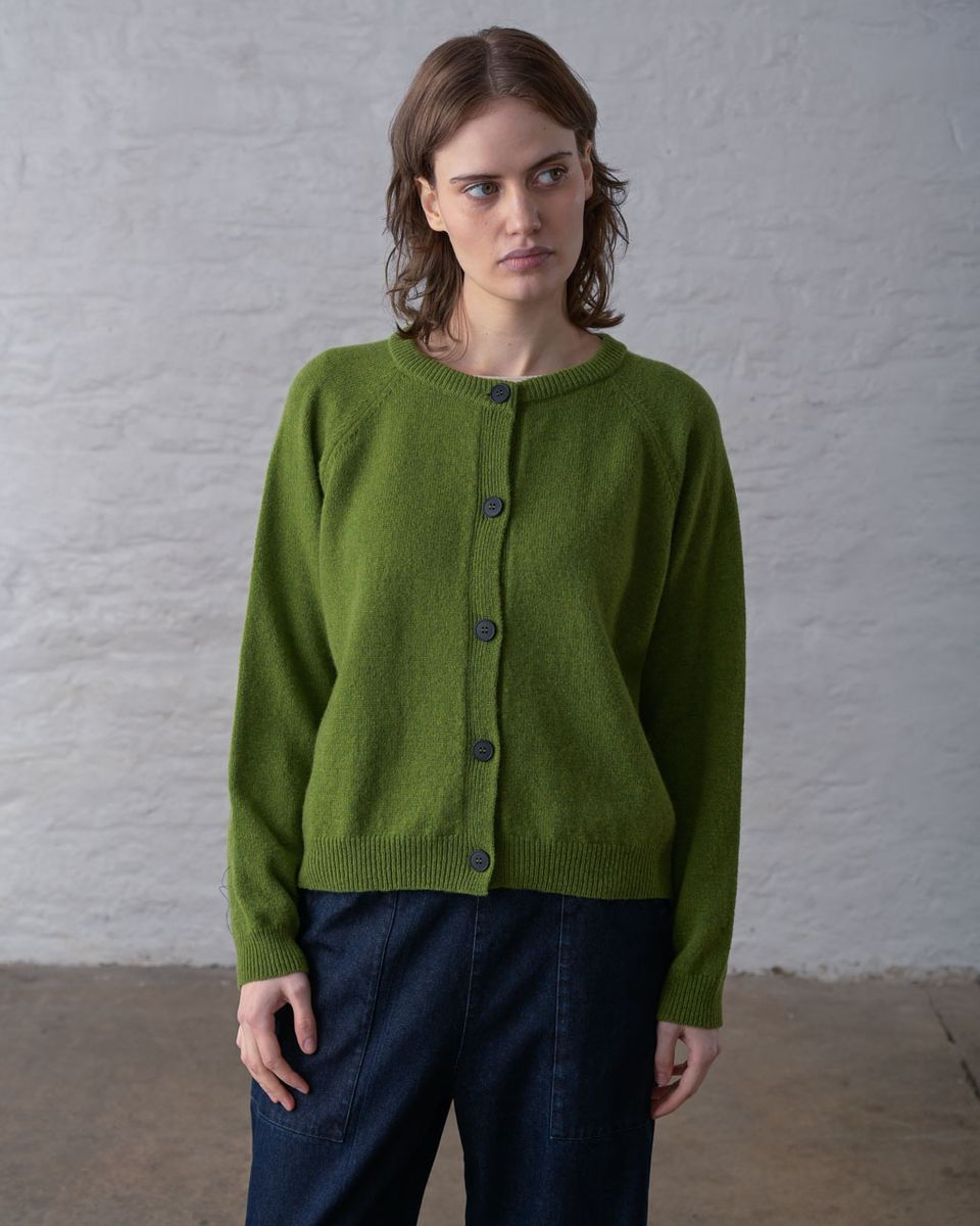 Front view of a woman wearing the BIBICO Lottie Cardigan in Moss buttoned closed, showing the ribbed round neckline, full button front with dark imitation corozo buttons, raglan sleeves, ribbed cuffs, and relaxed hip-length silhouette in midweight moss green lambswool, styled with dark navy wide-leg trousers against a whitewashed brick wall
