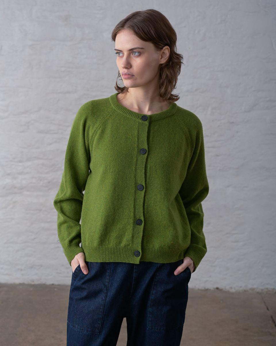 Three-quarter front view of a woman wearing the BIBICO Lottie Cardigan in Moss with hands in trouser pockets, showing the ribbed round neckline, dark imitation corozo buttons, raglan sleeves, ribbed hem, and relaxed hip-length fit in midweight moss green lambswool, styled with dark navy wide-leg trousers against a whitewashed brick wall
