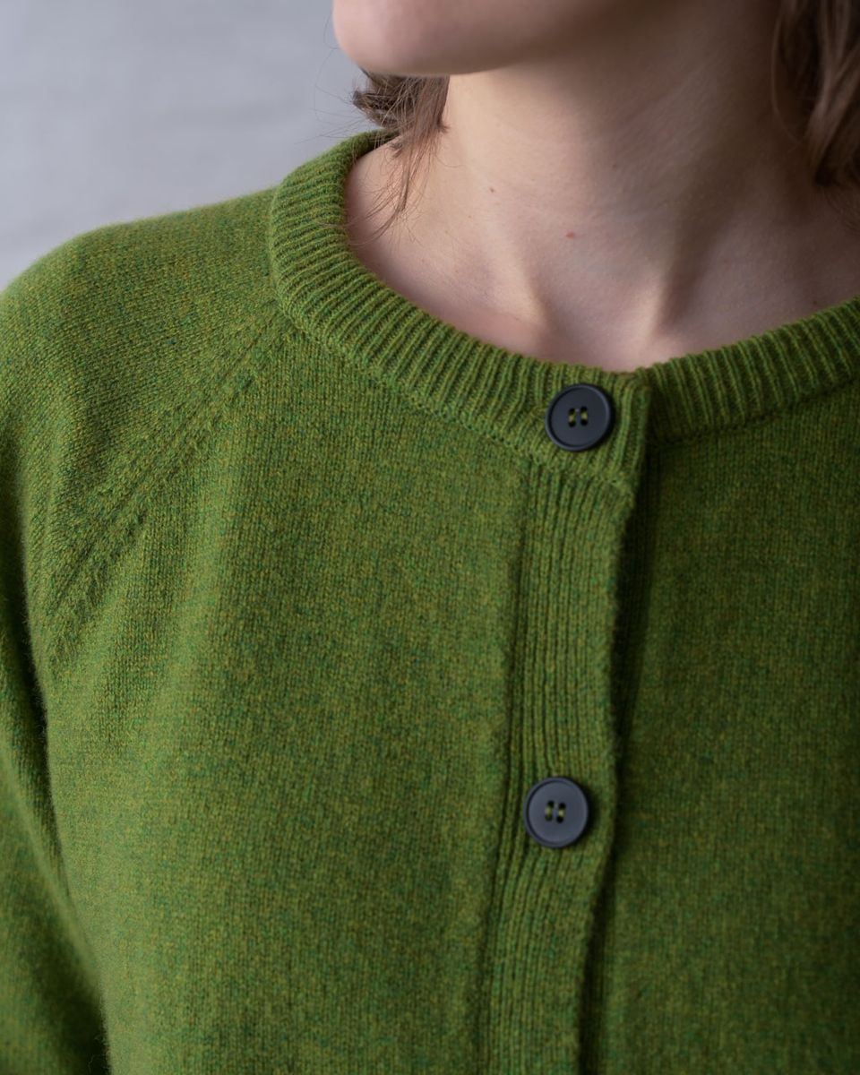 Close detail of the neckline of the BIBICO Lottie Cardigan in Moss, showing the fine ribbed round neck trim, dark imitation corozo buttons on the button placket, raglan sleeve join, and the soft texture of the midweight moss green lambswool
