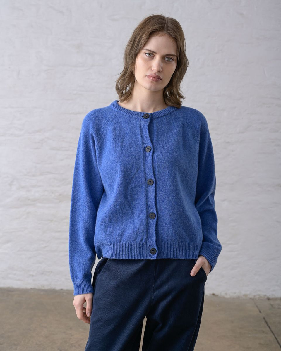 Front view of a woman wearing the BIBICO Lottie Cardigan in River buttoned closed, showing the ribbed round neckline, full button front with dark imitation corozo buttons, raglan sleeves, ribbed cuffs, and relaxed hip-length silhouette in midweight river blue lambswool, styled with dark navy wide-leg trousers against a whitewashed brick wall
