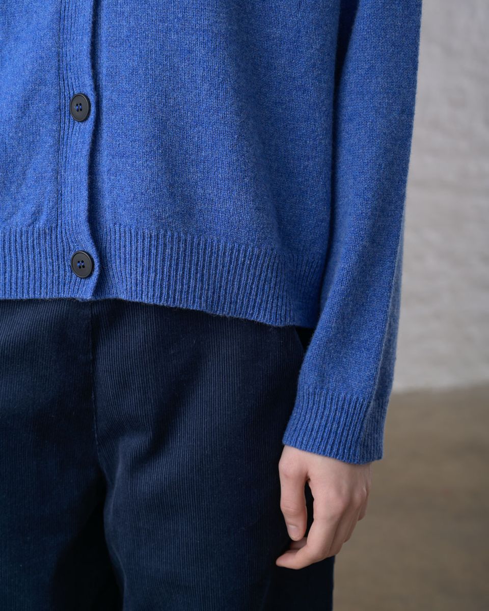Close detail of the hem and cuff of the BIBICO Lottie Cardigan in River, showing the ribbed hem, dark imitation corozo buttons at the button front, ribbed cuff, and the soft texture of the midweight river blue lambswool, styled over dark navy corduroy trousers
