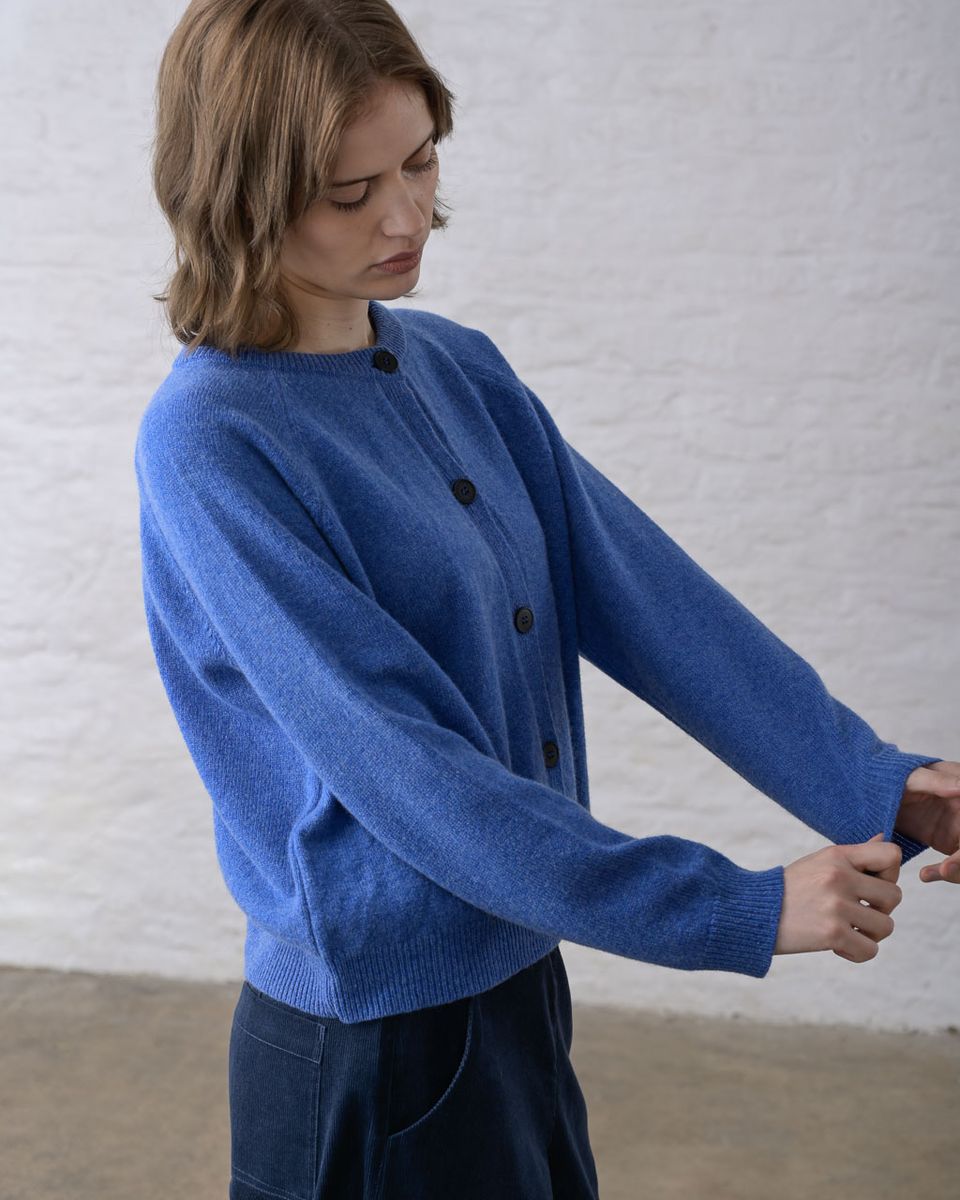 Side view of a woman adjusting the cuff of the BIBICO Lottie Cardigan in River, showing the raglan sleeve, ribbed cuff, button front with dark imitation corozo buttons, and relaxed fit in midweight river blue lambswool, styled with dark navy wide-leg trousers against a whitewashed brick wall
