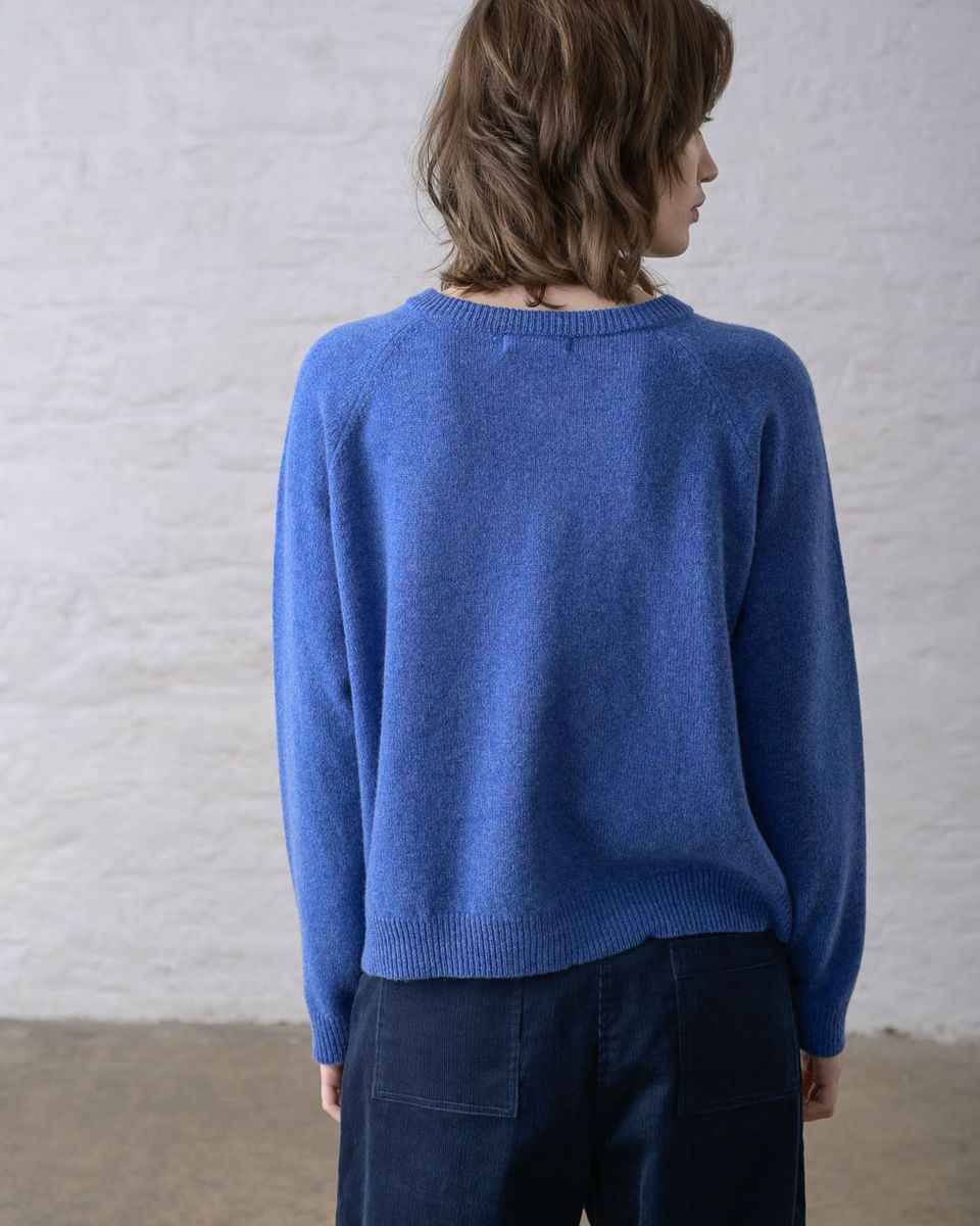Back view of a woman wearing the BIBICO Lottie Cardigan in River, showing the smooth fine knit body, ribbed round neckline, raglan sleeve seams, ribbed cuffs, and straight ribbed hem in midweight river blue lambswool, styled with dark navy wide-leg trousers against a whitewashed brick wall
